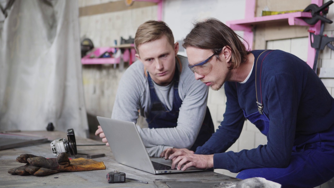 侧视图跟踪拍摄两个年轻的金属工人在笔记本电脑屏幕上讨论项目站在工作台周围的工具视频下载