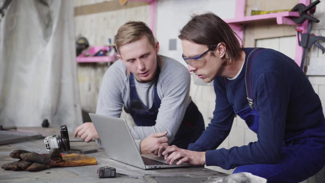 侧视图跟踪拍摄两个年轻的金属工人计划一起工作，使用笔记本电脑站在工作台周围的工具视频下载