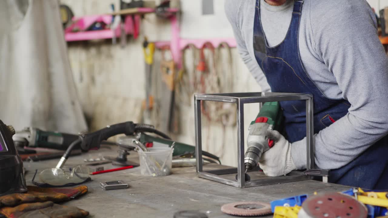 年轻的金属工人戴着防护手套，用电动螺丝刀将塑料零件固定在钢立方体上，站在车间工作台上制作灯，侧视跟踪镜头视频素材