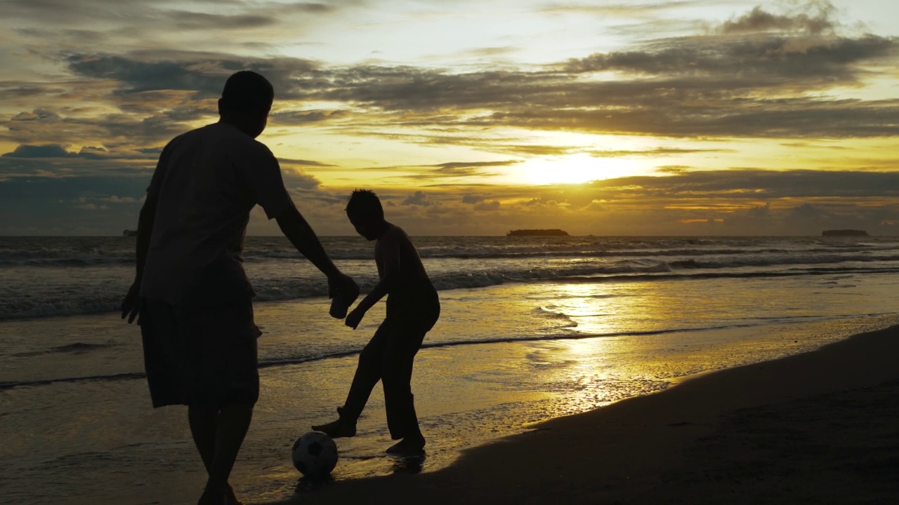 夕阳西下，幸福的父亲和儿子在海滩上踢足球。幸福的一家人在海滩上享受暑假。假日旅行的概念。假期视频素材