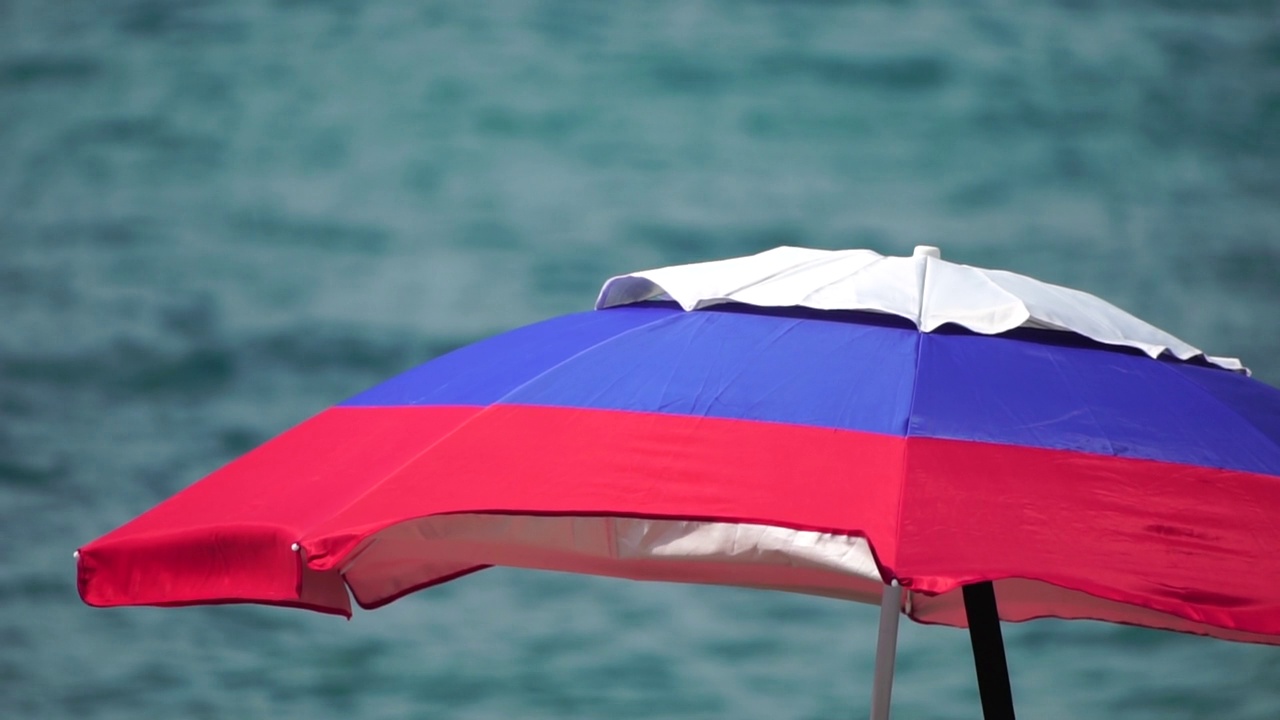 在一个阳光灿烂的日子里，在蓝色的大海上，白色、蓝色、红色的沙滩伞或遮阳伞。象征着假期、放松、夏天。海滩上立着五颜六色的遮阳伞。慢动作视频下载