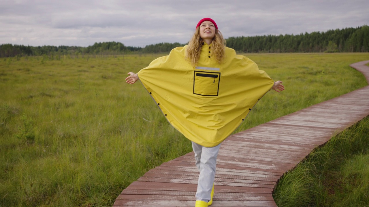 在多云的天气里，戴着红帽子和黄色雨披的漂亮女人在森林沼泽中间的一条木小路上散步视频下载