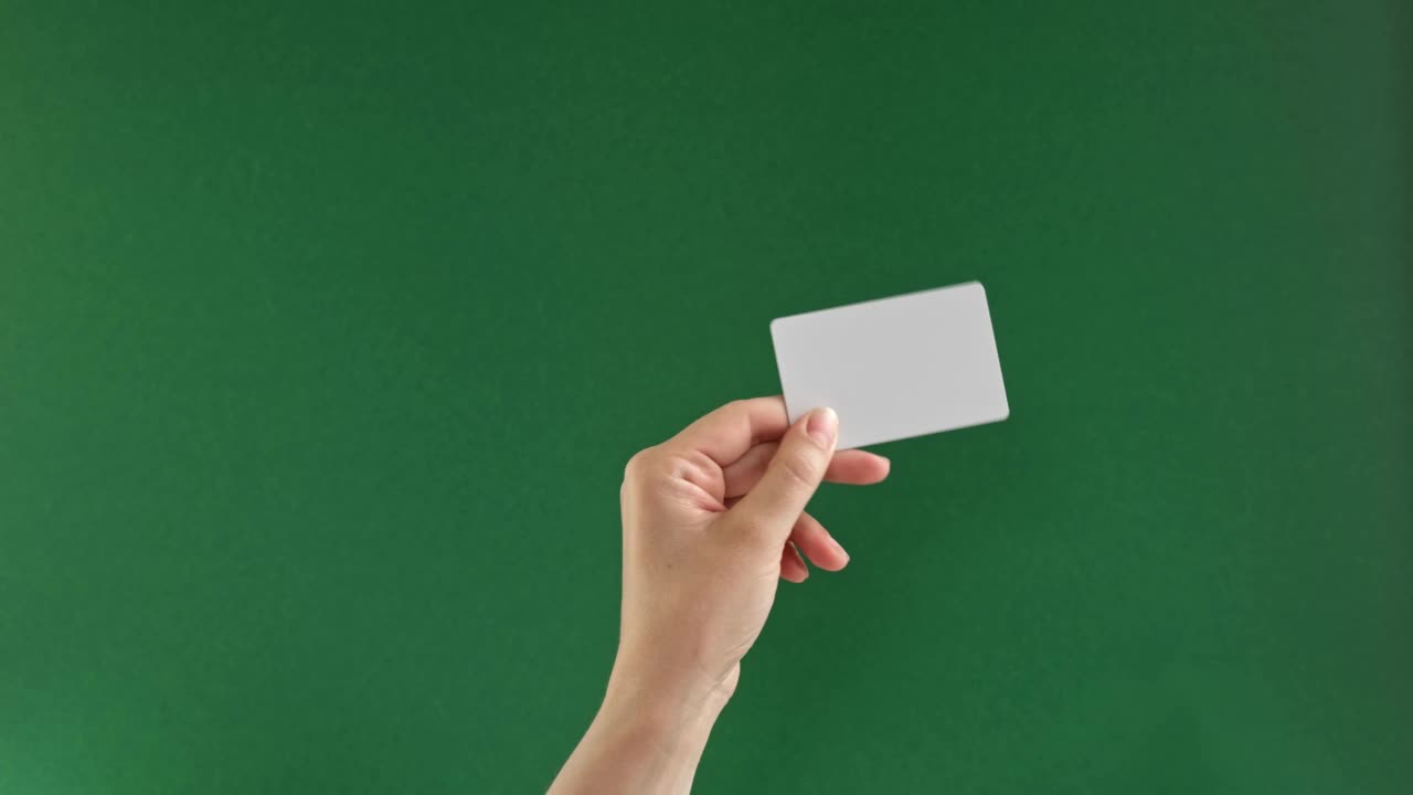 女性手握白色信用卡，绿色背景隔离。关闭了。购买圣诞礼物。色度键屏幕。女人的手与塑料礼品卡模型。广告的姿态。4 k视频下载