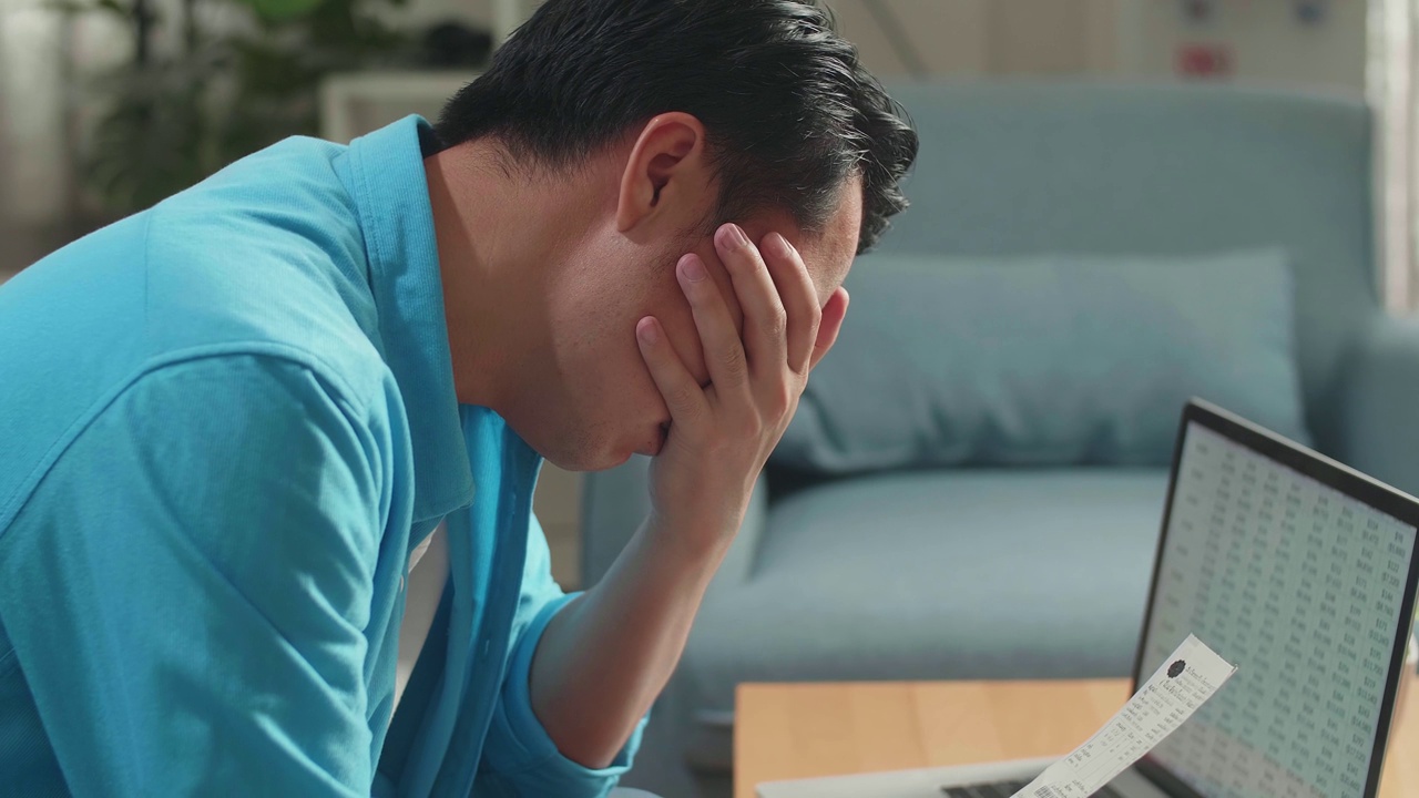 一个亚洲男人带着笔记本电脑看账单，因为财务问题而头痛视频下载