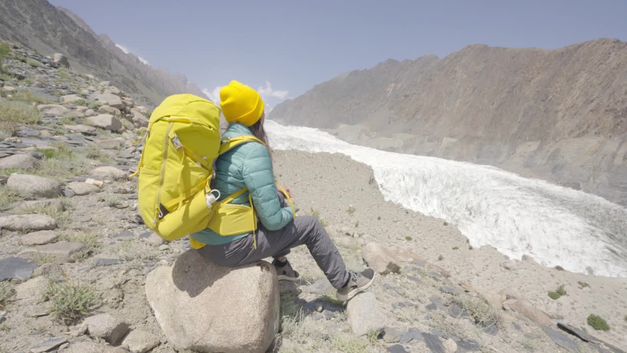 自由旅行者的后景年轻的女人穿着冬衣背着黄色的背包坐在岩石上看早晨的冰川。体验旅游的概念。与α香奈儿。视频素材