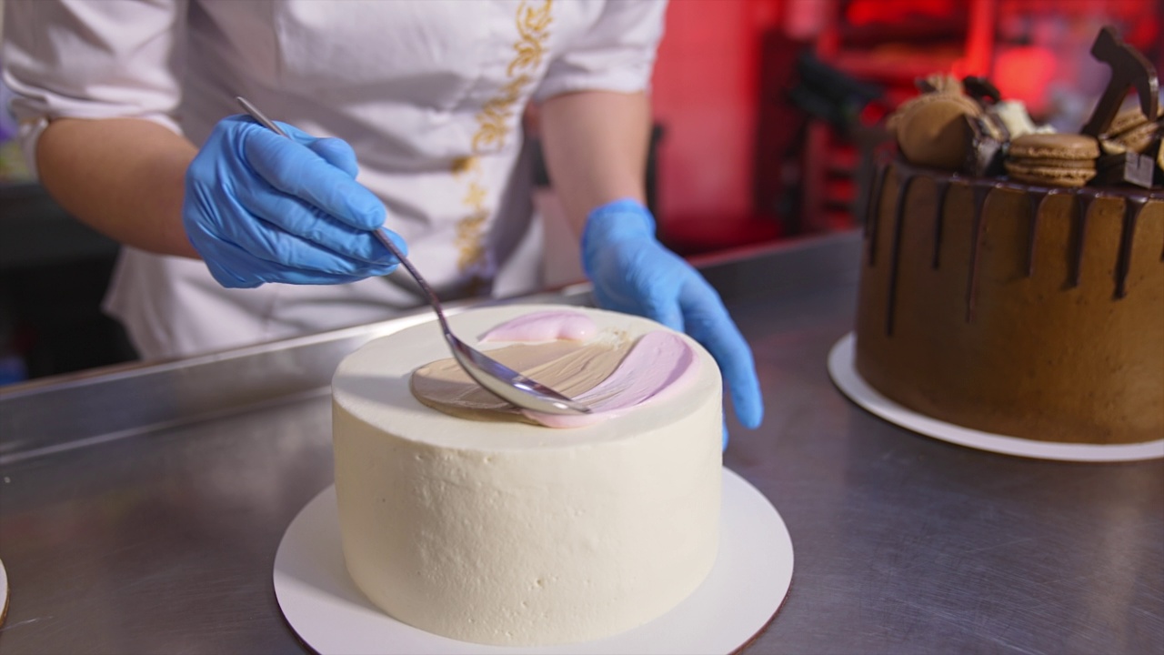 糕点师用勺子把白奶油涂在白蛋糕上。关闭了。背景桌上的巧克力蛋糕。视频下载