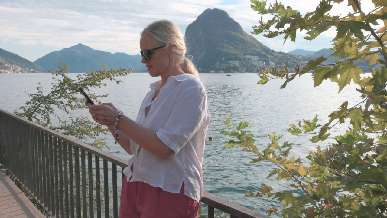 一名女子在俯瞰湖面的栏杆旁放松，使用智能手机视频下载