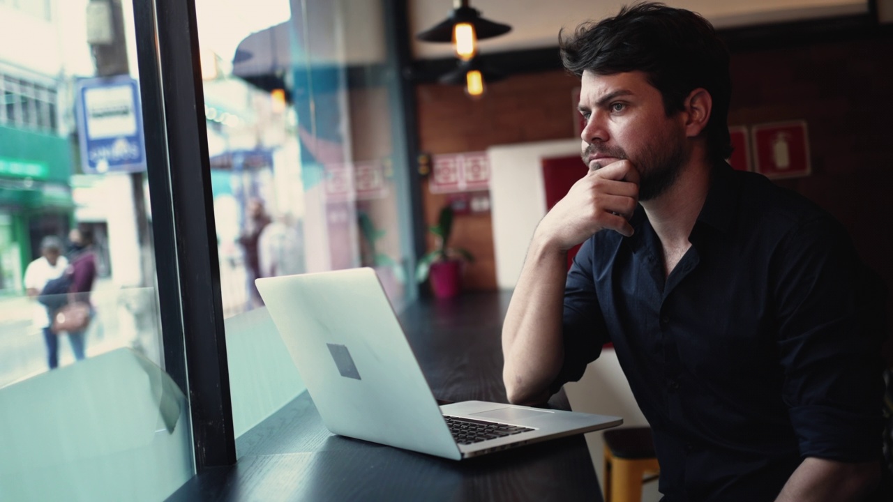 关心沉思的年轻企业家男子在笔记本电脑前坐在咖啡馆的窗口合作远程工作场所视频下载