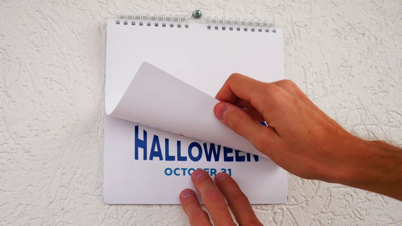 特写:一个男人的手撕下一张黑色的纸，上面写着“十月”，接着是另一张有万圣节大南瓜和日期的纸视频下载