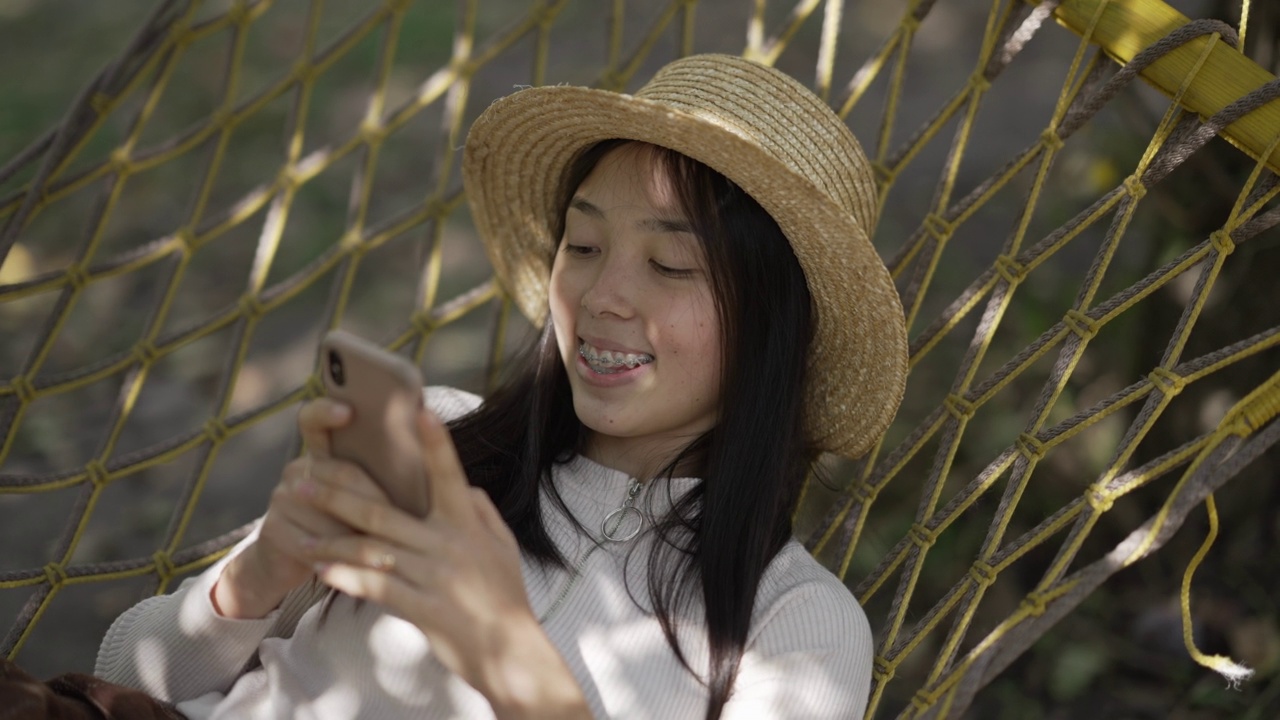 高角视角的快乐放松的亚洲女性在智能手机应用程序在线消息躺在户外吊床上。画像快乐无忧无虑苗条美丽的女士在草帽享受悠闲的公园。视频素材