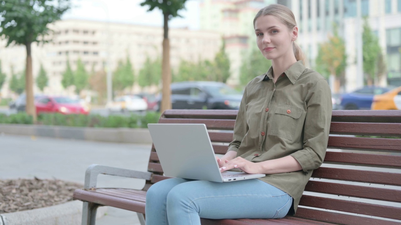 拿着笔记本电脑的妇女坐在户外长凳上对着镜头微笑视频素材