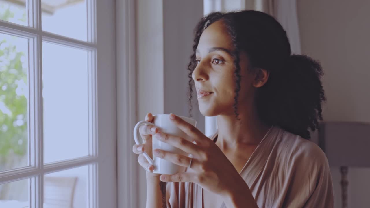 快乐的女人喝着一杯咖啡，看着房间窗外的早晨在家里。美丽的内容女性卷发思考和享受一杯茶。欣赏景色视频素材