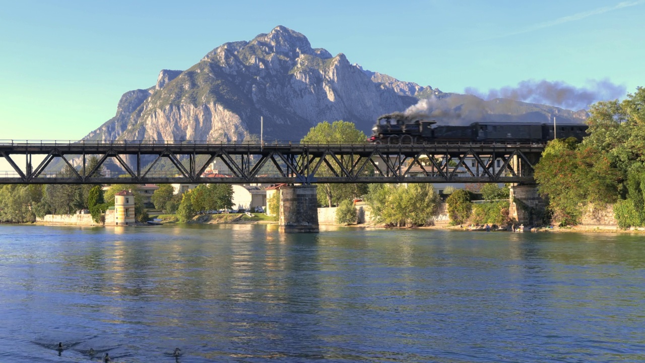 古老的蒸汽机车穿过河流上的古铁桥，背景是山脉。蒸汽火车视频下载