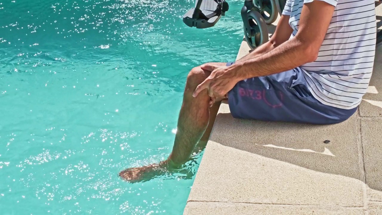 坐在轮椅旁边的残疾人，把脚放在泳池里，移动他的腿来激活血液循环视频下载