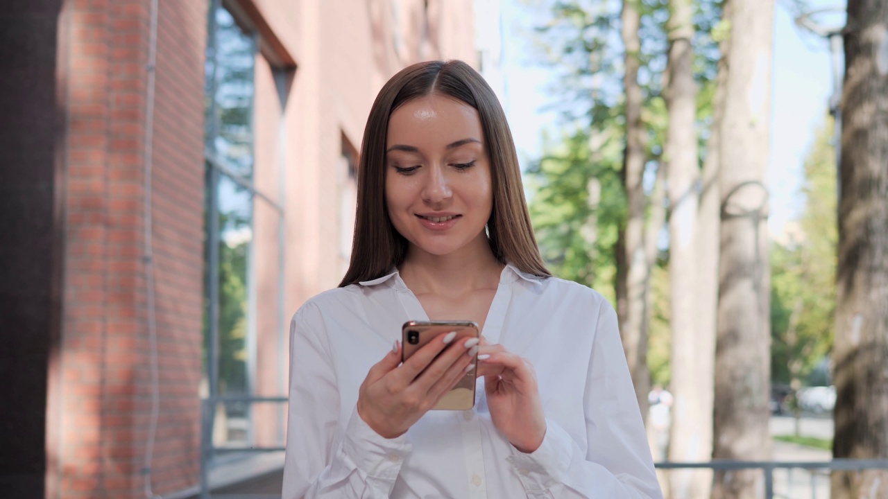 可爱的年轻女商人在白衬衫拿着智能手机写电子邮件在户外工作。夏天的早晨，聪明的女人站在公园现代建筑附近浏览互联网。业务人员。视频素材