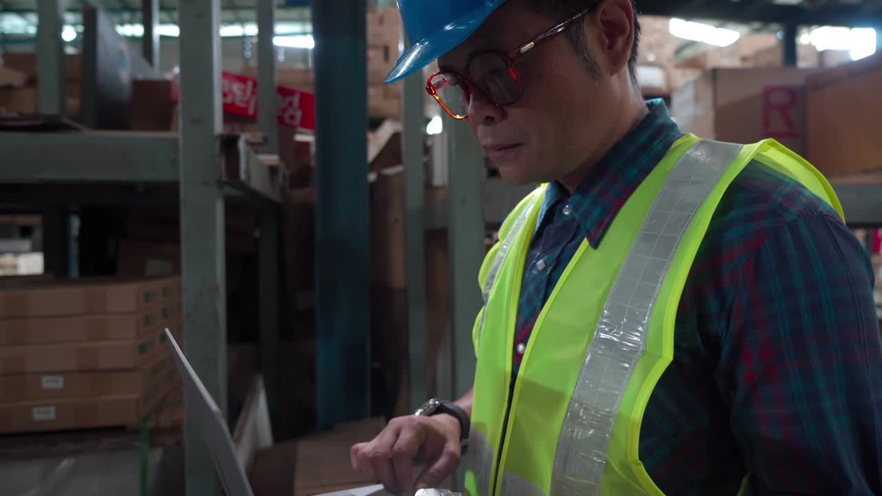 戴眼镜的亚洲男子站在仓库用笔记本电脑检查库存，男性工程师戴防护头盔和反光背心，工人控制工厂安排视频下载