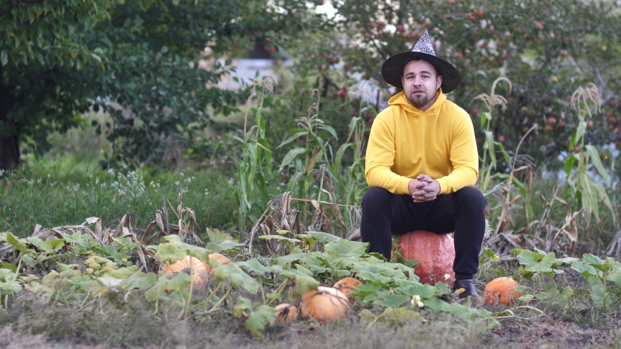 一个年轻的男性农民博主坐在南瓜田里的一个巨大的南瓜上，戴着女巫的帽子，对着镜头说话。蔬菜的影响视频素材