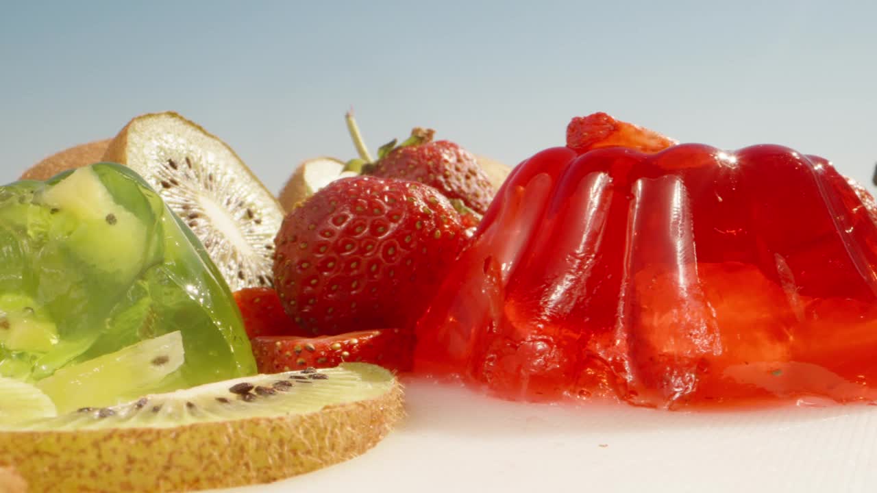 红绿果冻，里面有猕猴桃和草莓。我用勺子拿了一块红色的果冻。桌上有草莓和猕猴桃。滑块极端特写镜头。视频下载