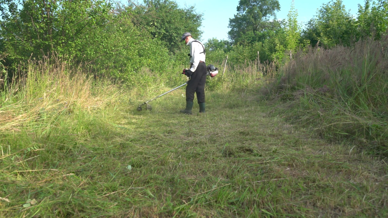 一个人用汽油镰刀割草。用割草机照料花园。视频下载