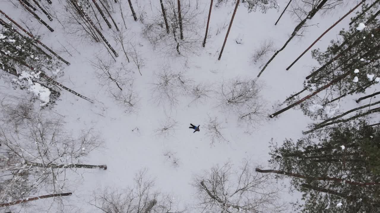 男孩躺在雪里，在雪林里的树中间，做雪天使。鸟瞰图。视频素材