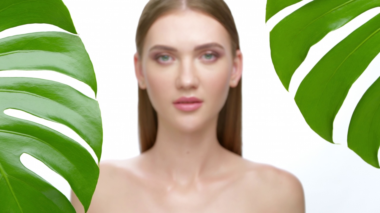 一个美丽的年轻女子的肖像与清洁和健康的皮肤在白色的背景，热带绿色的叶子在前景。视频下载