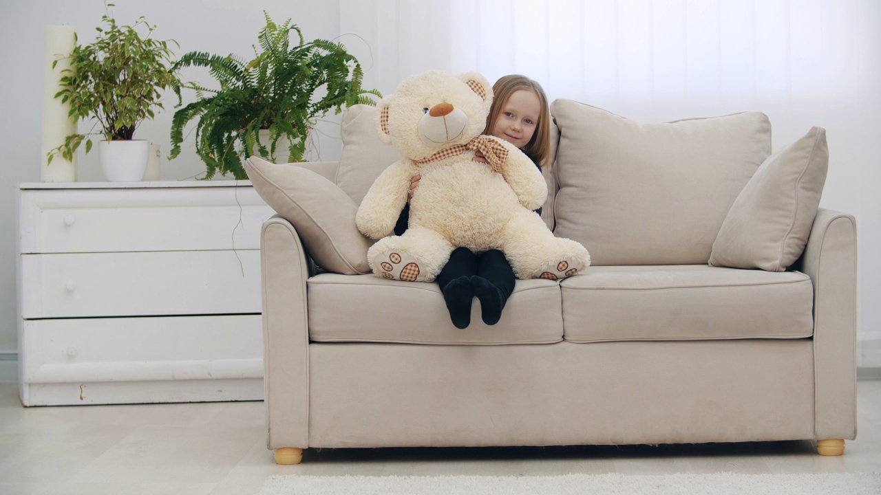 小女孩微笑着坐在沙发上，和她的玩具泰迪熊在4k慢动作视频中挥手问好。视频下载