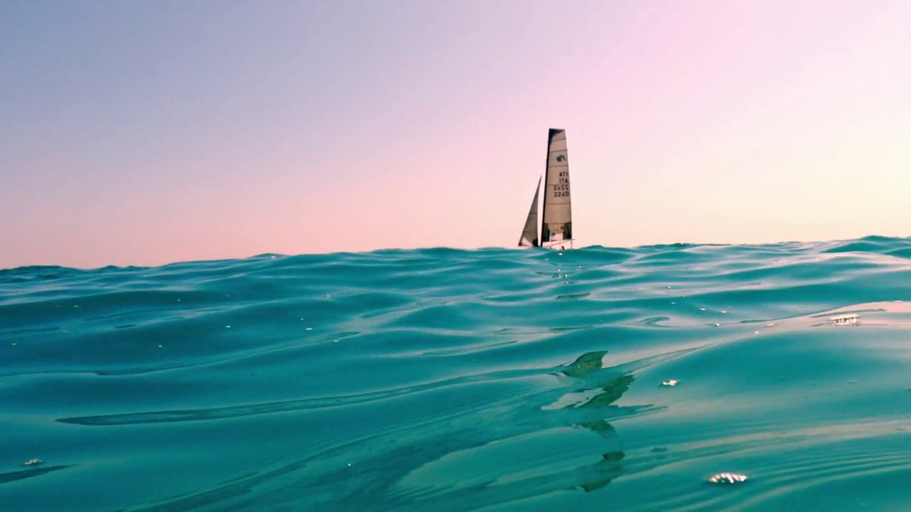 极低角度的海平面小帆船航行在平静开放的蓝绿色海水与粉红色的天空为背景视频素材
