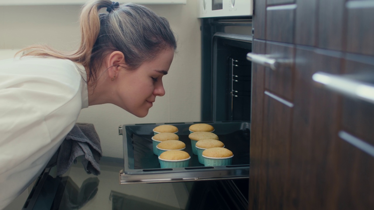 一个漂亮的女人从烤箱里拿出纸杯蛋糕，闻着它，在家里微笑视频素材