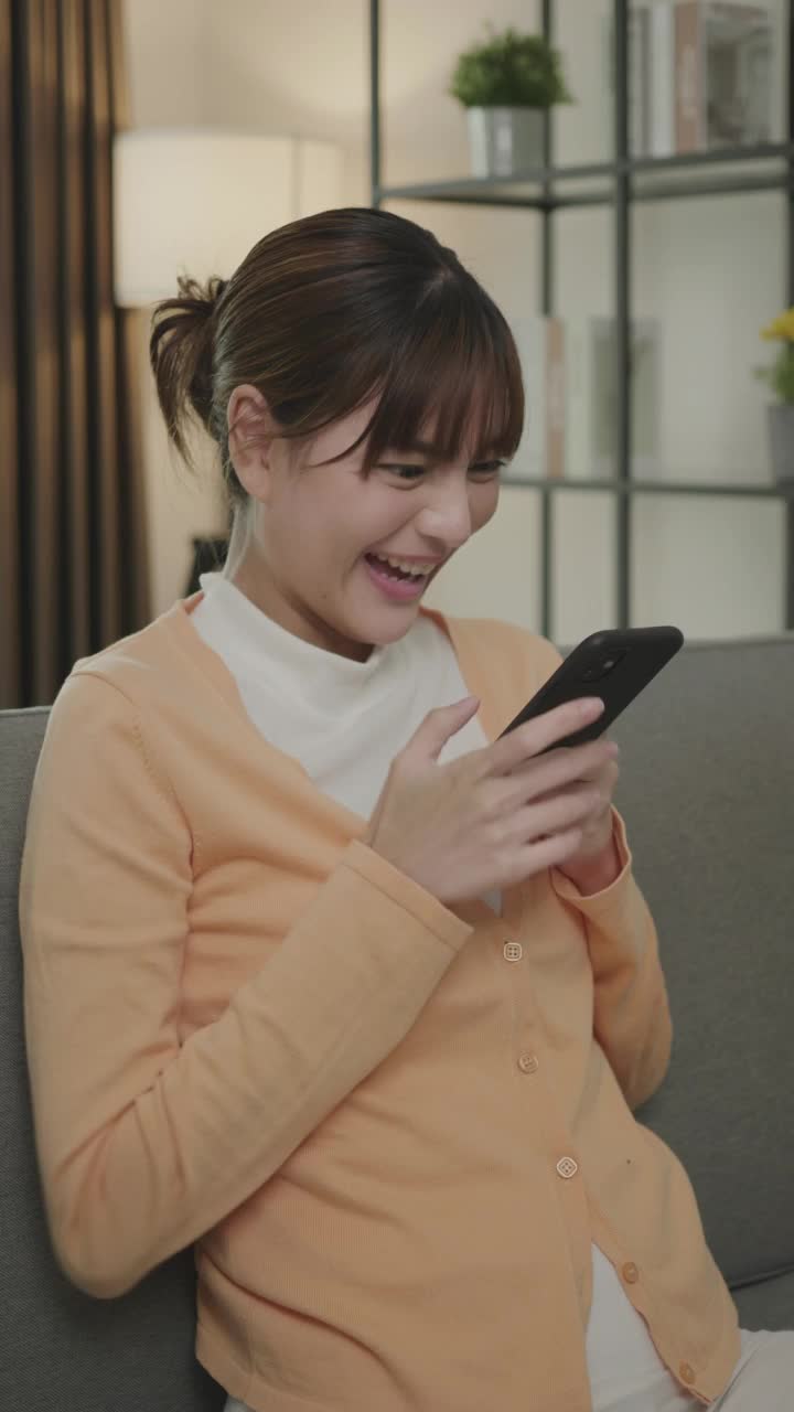 一位年轻的亚洲妇女在得到一份工作后高兴地笑着。一个女人坐在沙发上，她在家里玩智能手机。视频素材