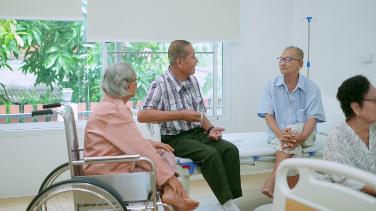 亚洲老年女性，戴着眼镜，坐在椅子上，喜欢和躺在养老院病人床上休息的朋友聊天，而其他老年朋友则在后面一起聊天，分享经验。视频下载
