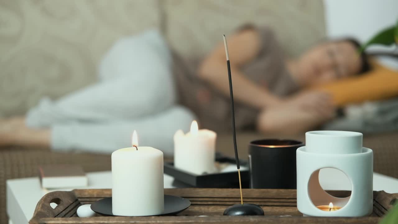 香薰蜡烛和香薰灯的特写。背景里的女人正躺在沙发上睡觉，很放松。抗压力和全面休息。心理健康的概念。视频下载