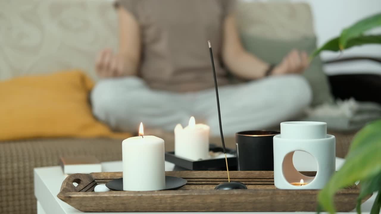 香薰蜡烛和香薰灯的特写。背景中，女子正坐在家里的沙发上，以莲花姿冥想。抗压力和全面休息。心理健康的概念。视频下载