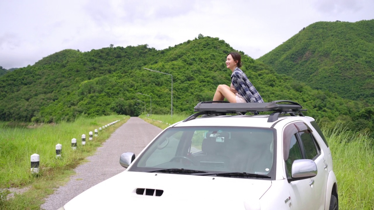 4K亚洲女子坐在汽车车顶架，举起手臂，而旅行在暑假自驾游。视频下载