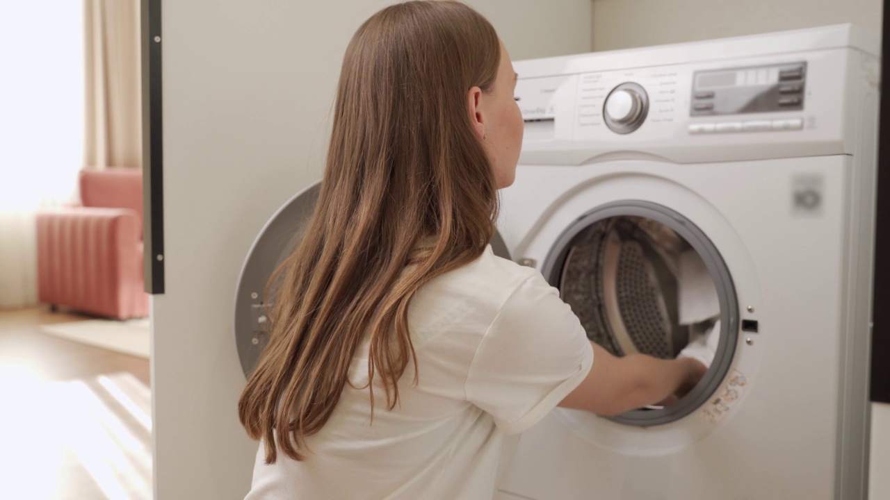 年轻友好的女人留着长发，穿着一件白色t恤，把篮子里的脏衣服放进洗衣房的白色洗衣机里。视频素材