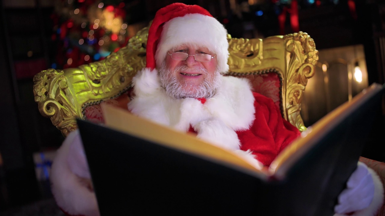 善良的老圣诞老人在他的房子里，挨着壁炉和装饰好的圣诞树，一边看书一边笑视频下载