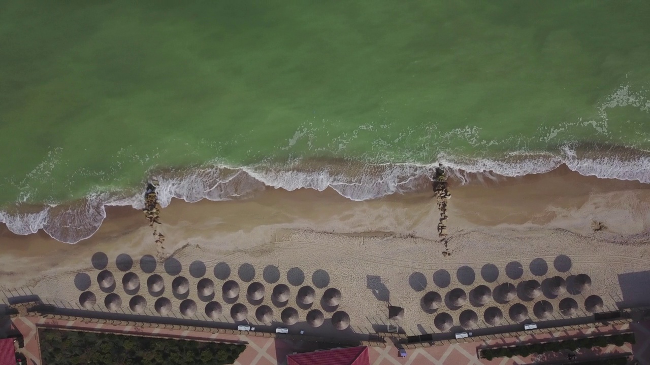 海滩上草伞的鸟瞰图。海浪冲刷着沙滩。一个豪华度假胜地的宁静海岸线。视频素材