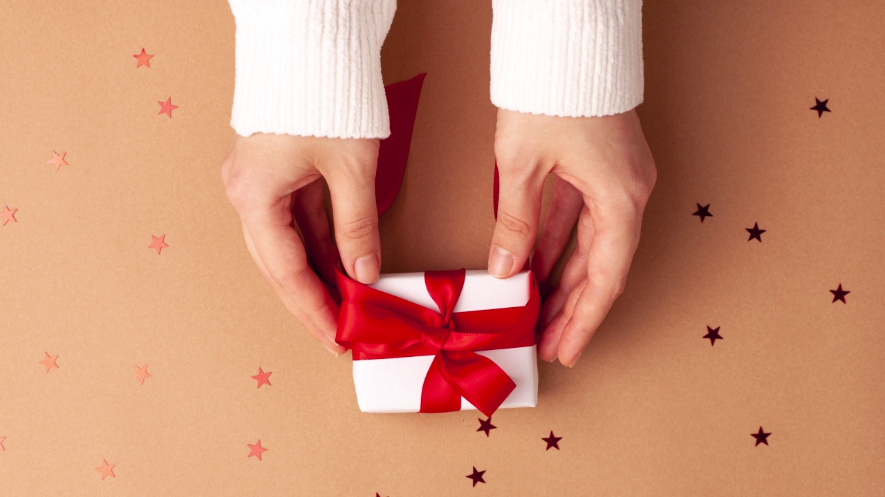 穿着白色毛衣的人手拿走了一个白色的纸礼物，上面有一个棕色背景的红色缎带蝴蝶结和红色的星星形状。定格动画圣诞假期概念平铺。视频下载