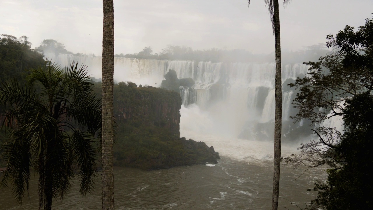 宽阔的白色瀑布透过两棵棕榈树，雾气从水中升起。视频素材