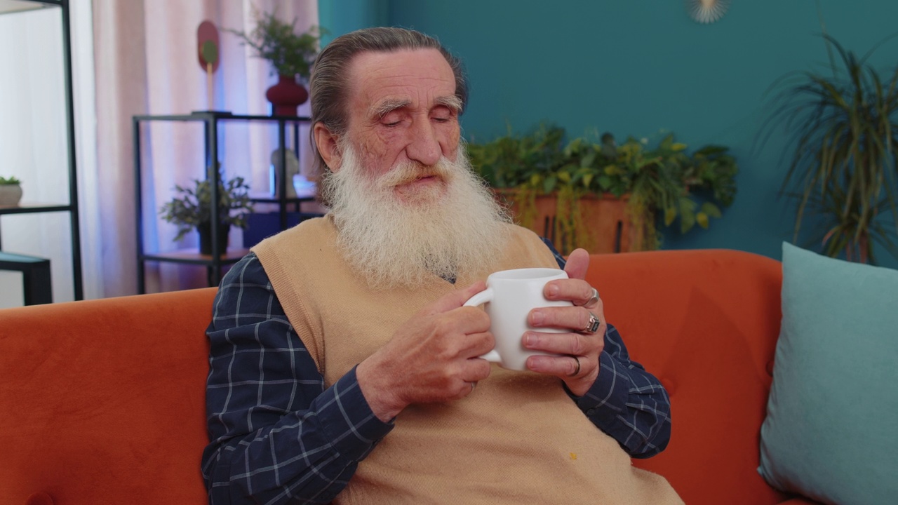 迷人的面带微笑的老爷爷坐在家里的沙发上喝着一杯咖啡或花草茶视频素材