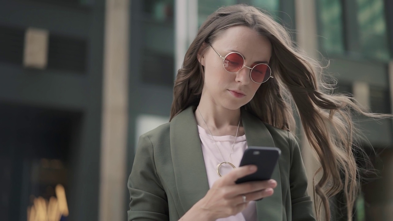 站在城市街道上，戴着红色太阳镜的年轻女子正在看她的智能手机视频素材
