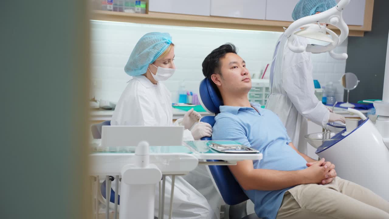 白人牙医在牙科诊所为年轻病人检查牙齿。帅哥躺在牙科椅上，在医院预约就诊时接受女医生的牙科治疗。视频下载