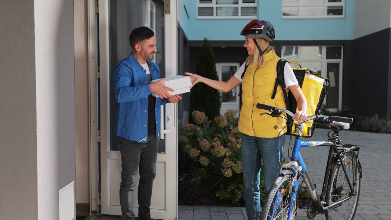 一名英俊的男子为骑着自行车的黄色保暖背包的披萨外卖女士开门，并收到了装着披萨的纸板箱。食品配送的概念。视频素材