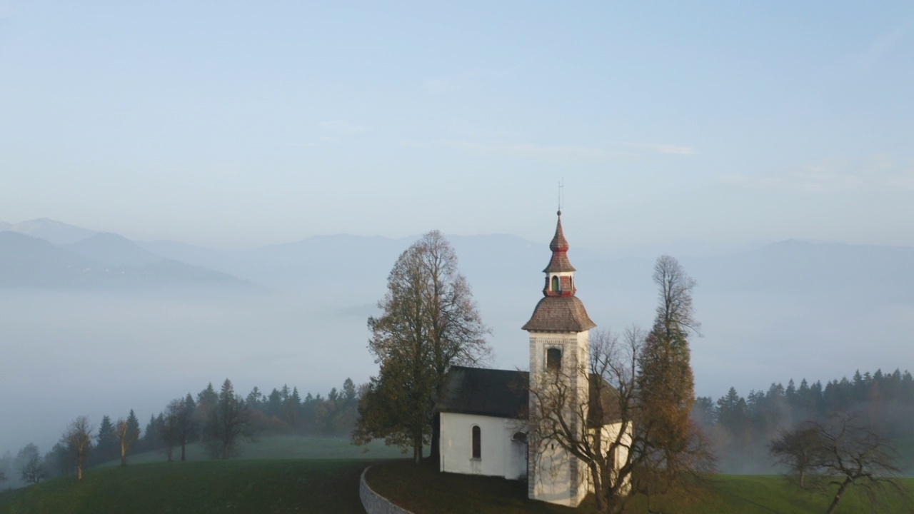 拂晓时分，在斯洛文尼亚的山上，飞过一座美丽的小教堂，雾气缭绕。美丽的秋天雾蒙蒙的风景在日出俯视图视频素材