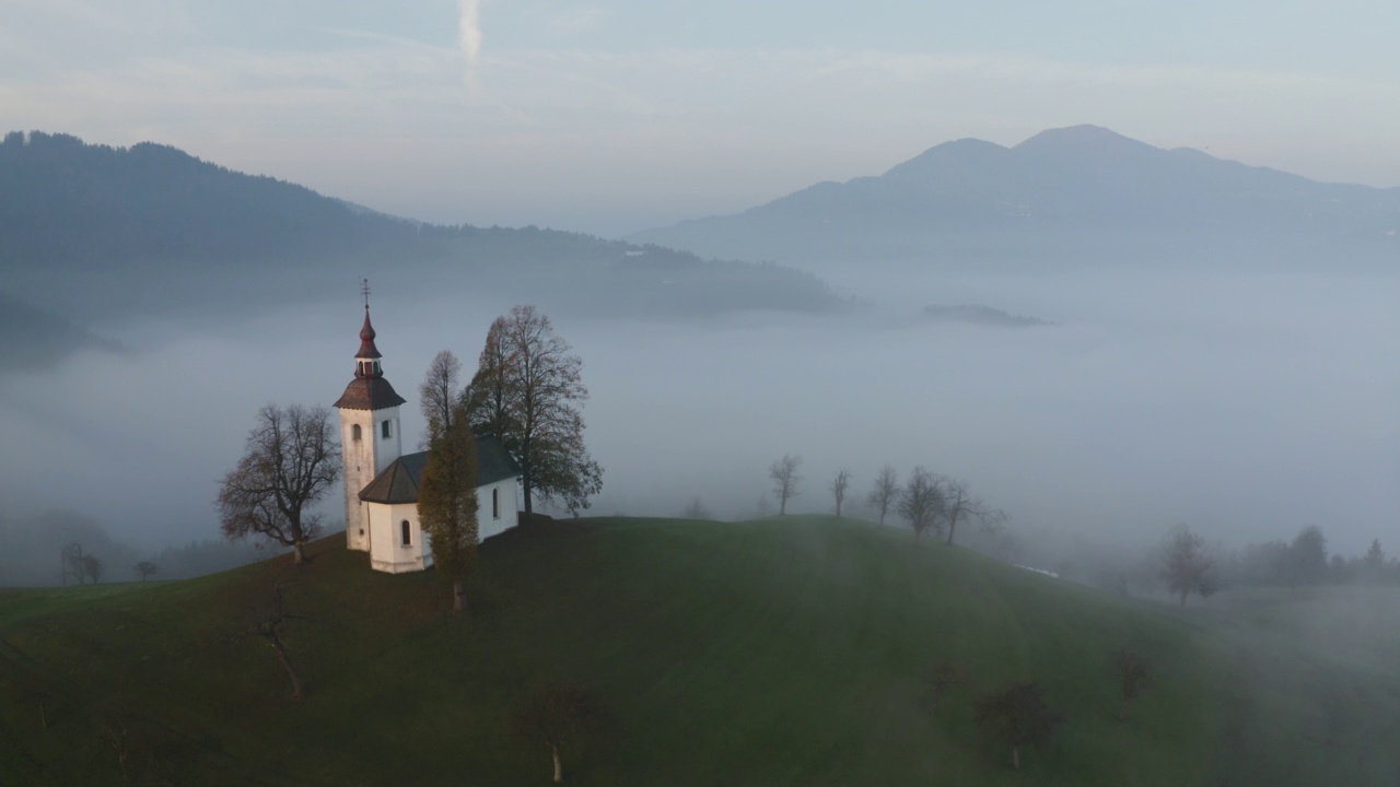 在日出俯视图有雾的山区上空飞行。无人机在黎明时分拍摄的斯洛文尼亚山上美丽的小教堂。美丽的秋雾风景视频素材