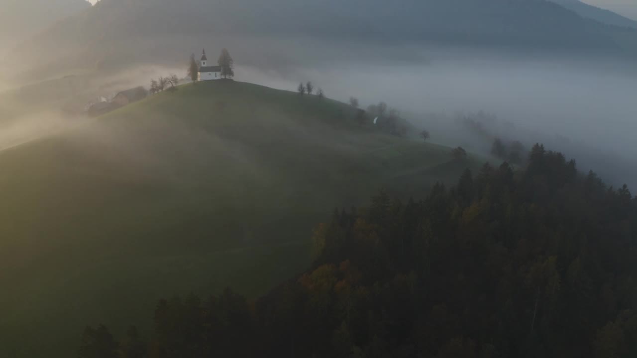 无人机在黎明时分拍摄的斯洛文尼亚山上美丽的小教堂。在日出俯视图有雾的山区上空飞行。美丽的秋雾风景视频素材