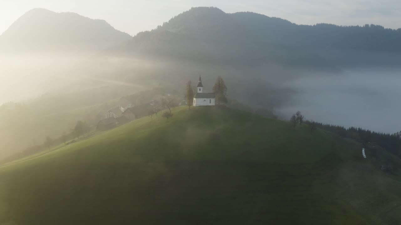 无人机在黎明时分拍摄的斯洛文尼亚山上美丽的小教堂。在日出俯视图有雾的山区上空飞行。美丽的秋雾风景视频素材