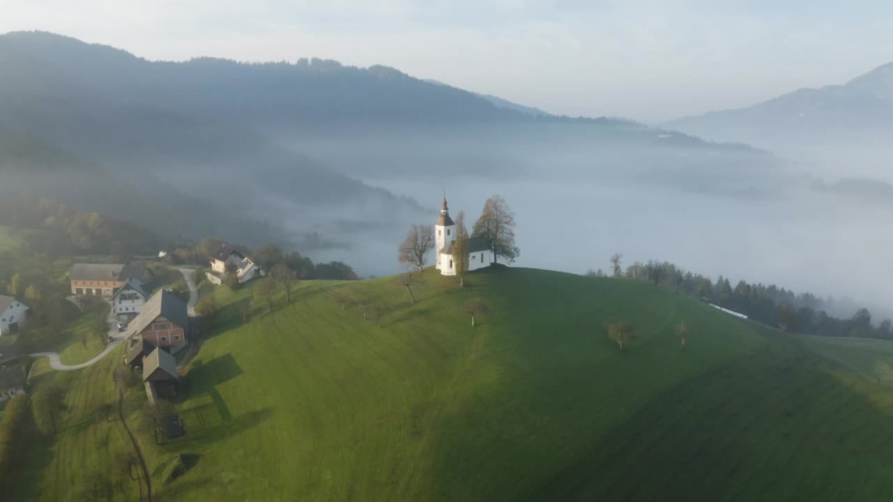 无人机在黎明时分拍摄的斯洛文尼亚山上美丽的小教堂。在日出俯视图有雾的山区上空飞行。美丽的秋雾风景视频下载