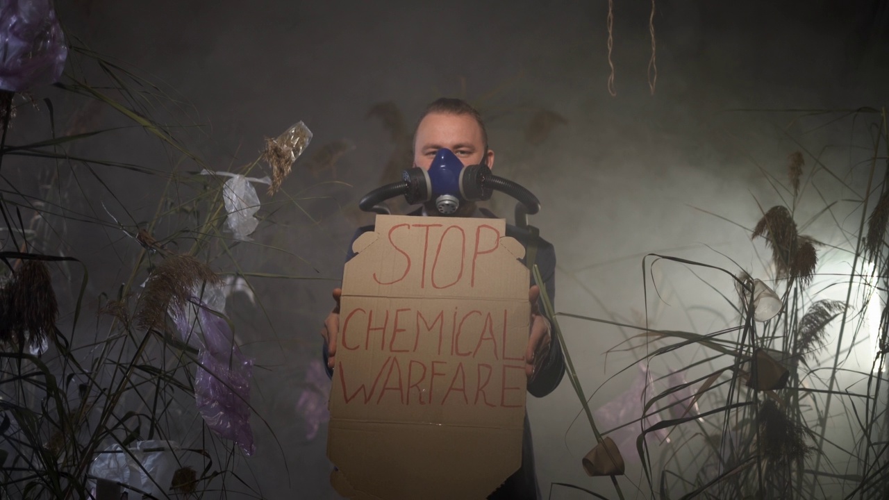 一名身穿夹克的男子站在烟雾弥漫的环境中，手里拿着禁止化学战的牌子。氧气罐与呼吸面罩相连。干草地上的塑料垃圾。生态灾难的概念视频素材