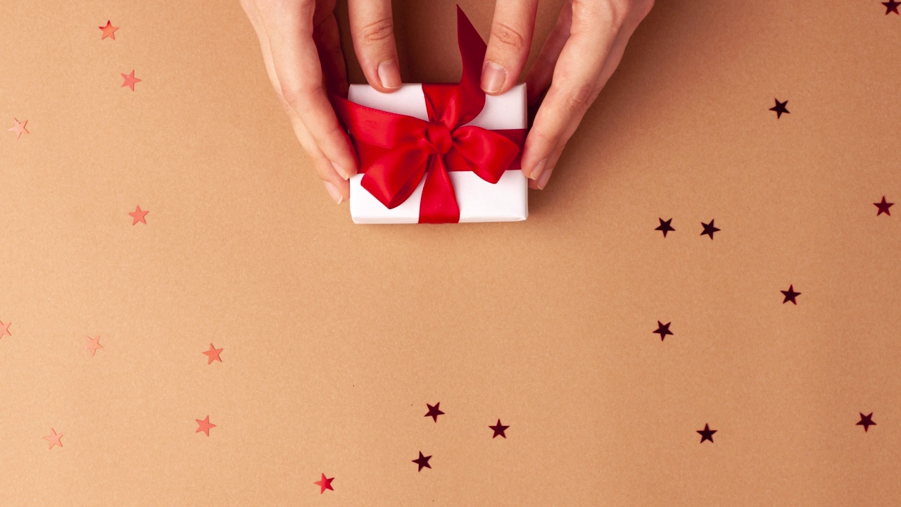 人类的手在白色的毛衣拿白色的纸礼物与红色缎带蝴蝶结在棕色的背景与红色的星星形状。定格动画圣诞假期概念平铺。视频下载