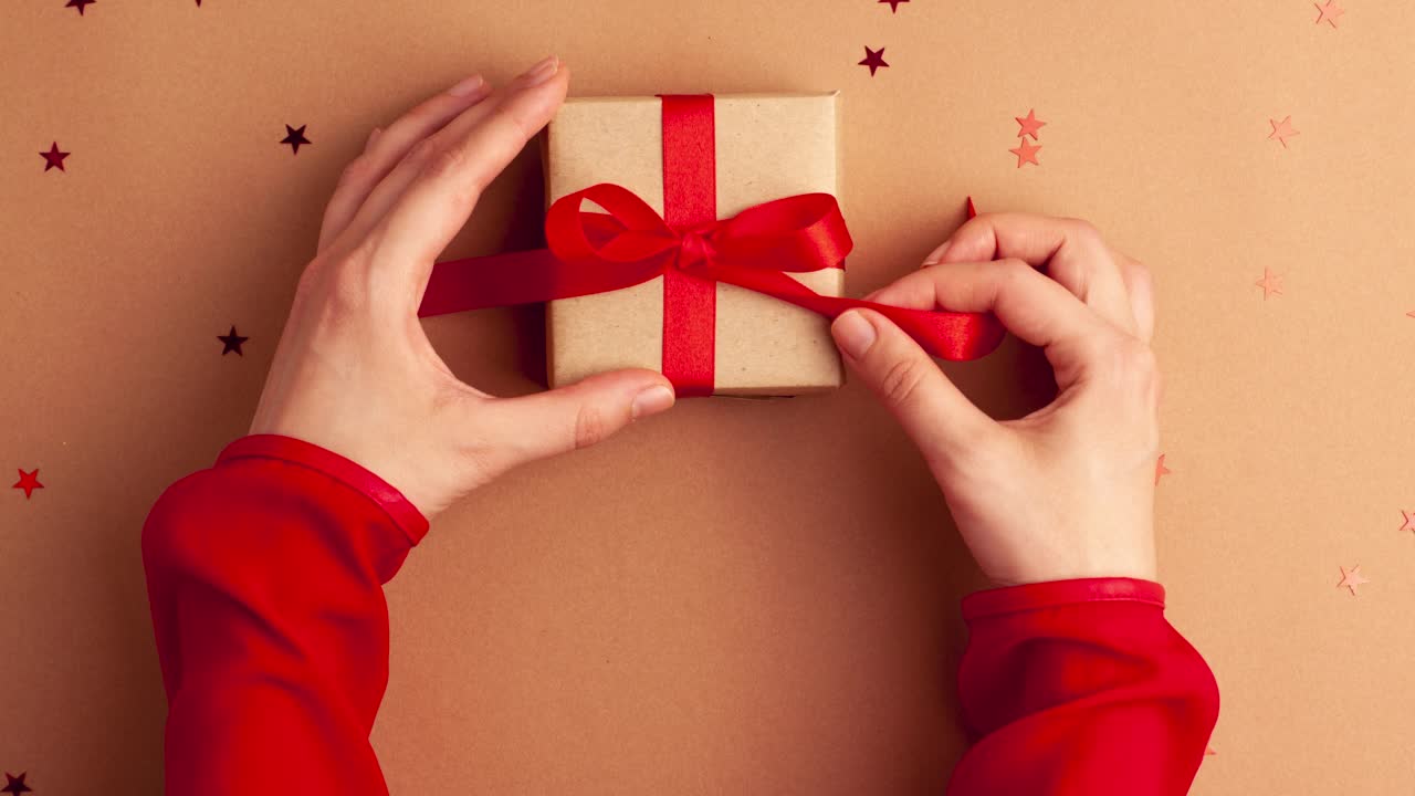 人手与红色袖子解开棕色纸礼物与红色缎带蝴蝶结在棕色背景与红色星星形状。定格动画圣诞假期概念平铺视频下载